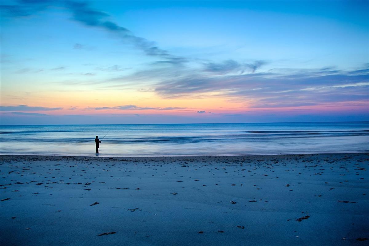 Person står på ei strand og fiskar i solnedgang. - Klikk for stort bilete