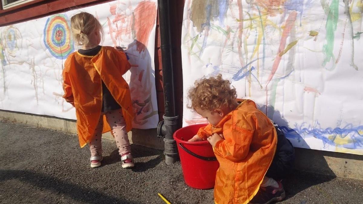 To barnehagebarn i oransje kitlar lagar veggmåleri. - Klikk for stort bilete